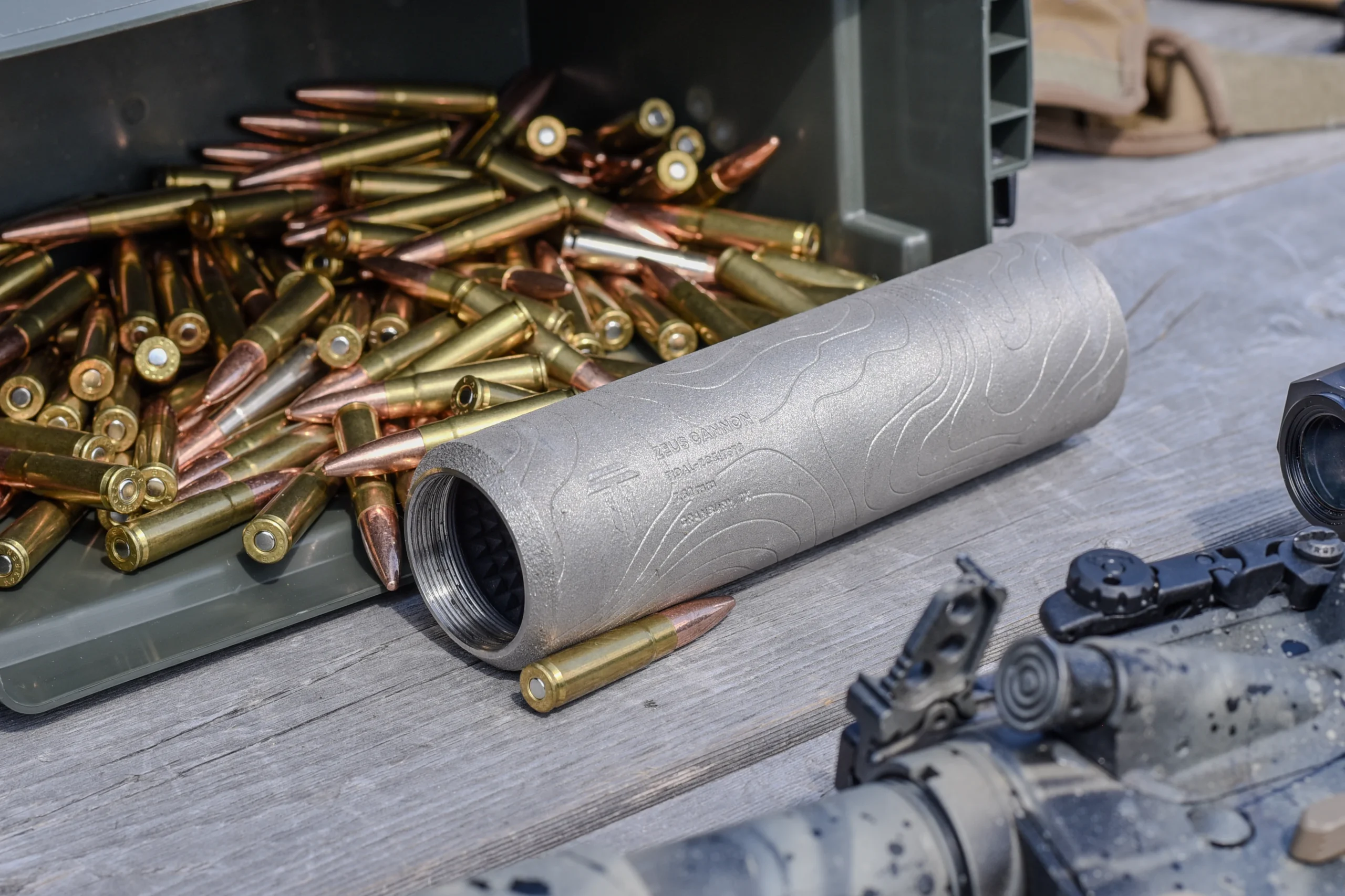 Raw titanium suppressor tube beside a spilled can of 7.62 brass cartridges on a weathered bench